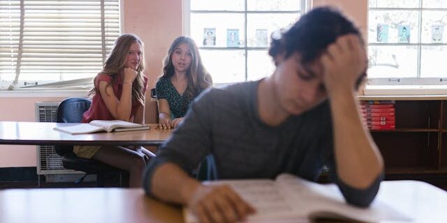 teen-2 boy in classroom upset and 2 girls behind him talking about him.