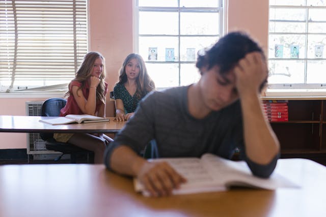 boy in classroom upset and 2 girls behind him talking about him.