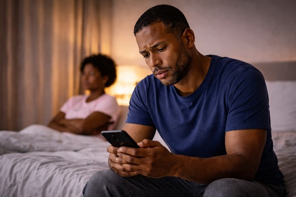 man sitting on the bed texting while his wife sits in the background with arms folded.